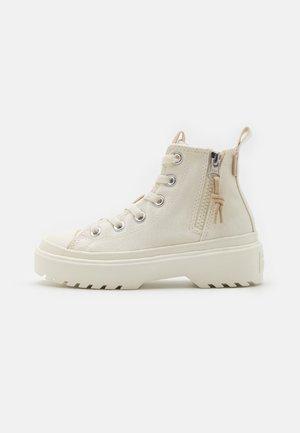Botas de tobillo beige hechas de lona, con ojales de metal, punta redonda, cremallera lateral, tirador y suela de goma texturizada con un patrón de relieve.