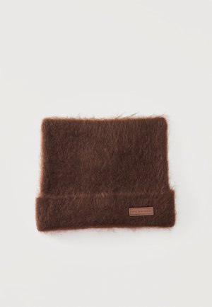 Pièce tricotée rectangulaire brune et duveteuse, avec un bord inférieur replié et une petite étiquette nominative, présentée sur un fond blanc.