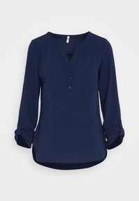 Blouse bleu marine à manches longues avec manches retroussables et patte de boutonnage à trois boutons, ourlet légèrement arrondi sur fond uni clair.