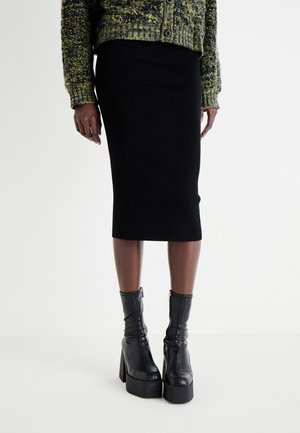 Persona con una falda lápiz midi negra, un suéter verde texturizado con botones y botas negras de plataforma hasta el tobillo con tacones apilados.