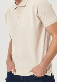 Beige pikétröja i mjuk bomull, med klassisk krage, två knappar och ribbad mudd. En avslappnad passform med sidslitningar.