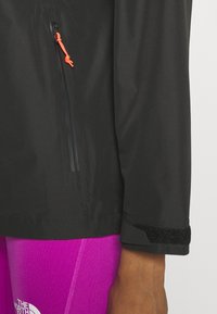 Giacca leggera nera con un toggle arancione regolabile, dotata di una tasca con zip, dettagli in velcro sui polsini e una superficie liscia.