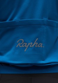 Κοντινό πλάνο μπλε υφάσματος με το λογότυπο "Rapha." τυπωμένο πάνω από οριζόντια ραφή, πιθανώς μέρος της πίσω τσέπης ενός ποδηλατικού τζέρσεϊ.