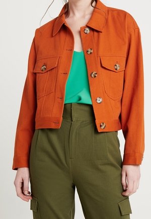 Kvinde iført en brændt orange knappet jakke over en blågrøn top med højttaljede olivengrønne cargobukser.