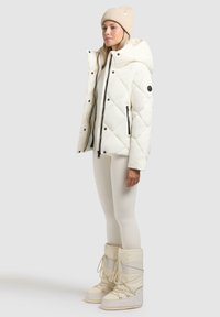 Witte gewatteerde pufferjack met een capuchon, zwarte accenten en volledige rits, gecombineerd met een beige gebreide beanie en witte snowboots.