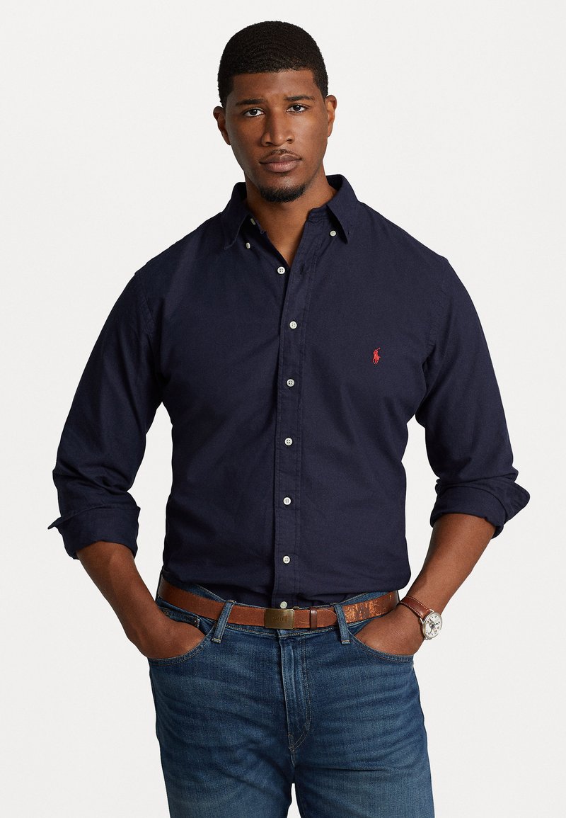 Chemise bleu marine à boutons en coton avec des manches longues ; logo rouge sur la poitrine. Accompagnée d'un jean bleu et d'une ceinture marron ; montre argentée au poignet.