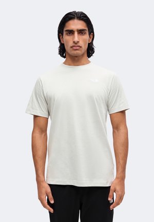 Homme aux cheveux noirs mi-longs portant une chemise légère unie à manches courtes et un pantalon foncé, debout devant un fond blanc.