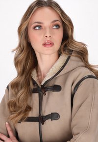 Beige hoodiejas met bruine leren sluitingen, contrasterende zwarte accenten en een zachte, gestructureerde bontvoering rond de kraag.
