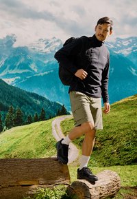 Un homme portant un pull en polaire zippé noir, un short kaki et des chaussures de randonnée noires, se tenant sur une bûche dans un cadre montagnard en extérieur.