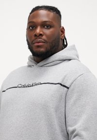 Szary sweatshirt z kapturem z czarnym logo Calvin Klein i poziomą paską. Miękka tekstura oraz luźny krój.