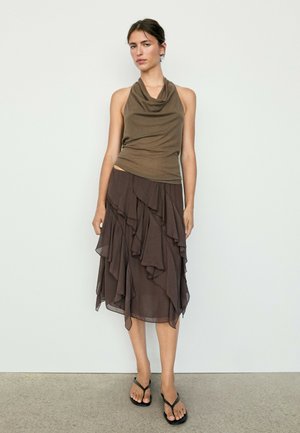 Femme portant un top sans manches col bénitier vert olive et une jupe asymétrique superposée marron foncé avec des sandales noires, debout contre un mur blanc.