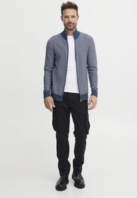 Cardigan de punto azul marino con puños y cuello de canalé, combinado con una camiseta blanca y pantalones cargo negros, que cuentan con múltiples bolsillos y un ajuste relajado.