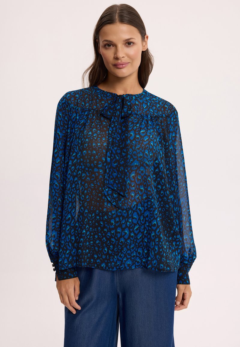 Kobieta ubrana w przejrzystą, niebiesko-czarną bluzkę w panterkę z długim rękawem i wiązaniem pod szyją oraz w granatowe, szerokie spodnie.