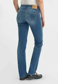 Jeans en denim bleu avec une taille mi-haute, coupe slim, deux poches arrière et une étiquette en cuir beige au-dessus de la taille à l'arrière. Texture délavée sur l'ensemble.