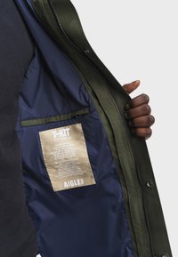 Zelená parka s námořnickě modrou podšívkou, která má nášivku s detaily o produktu a zipovou kapsu uvnitř.