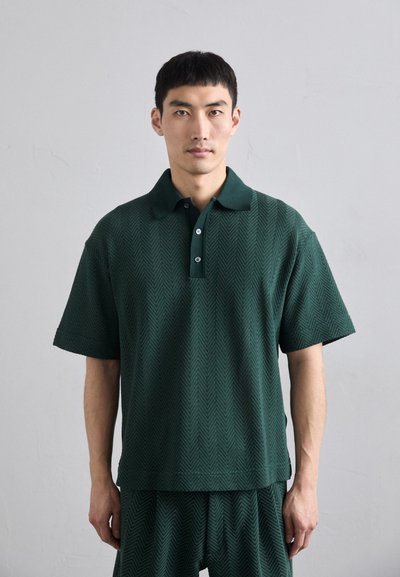 Grøn kortærmet polo lavet af tekstureret materiale med et herringbone-mønster, der har en krave og en tre-knaps lukning.