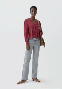 Frau in einer roten Bluse mit langen Ärmeln, hellgrauen Jeans, flachen Schuhen im Kuhflecken-Muster und einer braunen Schultertasche vor einem schlichten weißen Hintergrund.