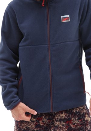 Chaqueta polar azul marino con cremallera frontal, detalles rojos y bolsillos laterales. Presenta un logo en parche en el pecho izquierdo. Textura suave.