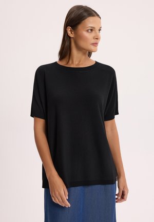 Kobieta o prostych brązowych włosach ubrana w luźny czarny top z krótkim rękawem i niebieską spódnicę, patrząca w prawo na jasnym, jednolitym tle.