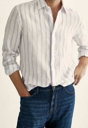 Mężczyzna w białej koszuli zapinanej na guziki z pionowymi jasnofioletowymi paskami oraz ciemnoniebieskich dżinsach, z rękami w kieszeniach.