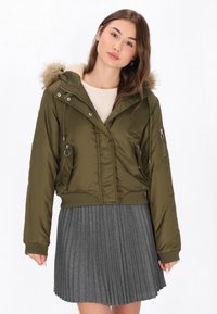 Giacca cropped verde oliva con cappuccio in pelliccia sintetica, tasche con zip e polsini a costine, abbinata a una gonna grigia plissettata.