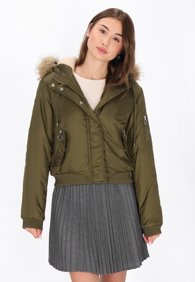 Giacca cropped verde oliva con cappuccio in pelliccia sintetica, tasche con zip e polsini a costine, abbinata a una gonna grigia plissettata.