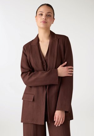 Giovane donna con capelli raccolti in uno chignon, indossa un blazer marrone strutturato e pantaloni abbinati, posando con un braccio incrociato sul petto.