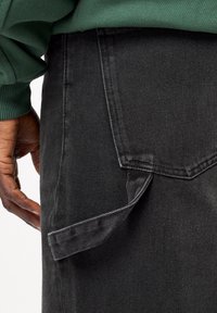 Schwarze Denim-Hose mit sichtbarer Gesäßtasche und einem Stoffschlaufen-Detail. Die Textur ist glatt mit leichtem Glanz, kombiniert mit einem Ärmel eines grünen Pullovers.