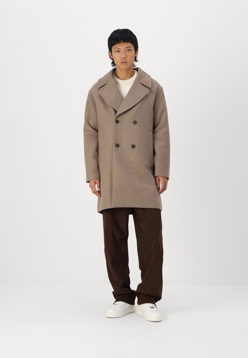 Palton din lână bej, cu un design cu guler dublu, rever mari, buzunare laterale și pantaloni din corduroy maro. Modelul poartă adidași albi.