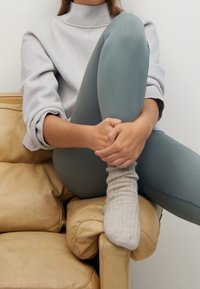 Persona sentada en una silla de cuero beige, vestida con un suéter gris claro, leggings de canalé verde azulado y calcetines grises, sosteniendo una rodilla cerca del pecho.