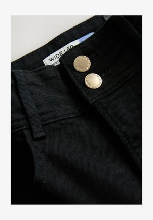 Prim-plan al taliei blugilor negri din denim, cu două nasturi metalici și etichetă albă vizibilă pe care scrie "Wide Leg".