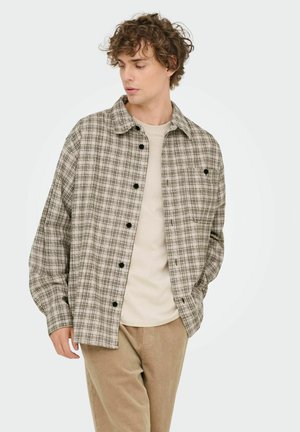 Geblokt shirt in beige en zwart, met een knoopsluiting aan de voorkant, een spread-kraag en lange mouwen. Ggedragen over een lichtgekleurde t-shirt.
