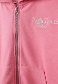 Ružičasti dukserica s patent-zatvaračem izrađena od mekanog pamuka, s natpisom "Paris Tennis Club" u svijetloj vezici. Ima središnji zatvarač i prednje džepove.