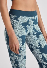 Femme portant des leggings bleus avec un motif floral paisley blanc, montrée de la taille jusqu'à mi-cuisse avec les mains posées sur les côtés.
