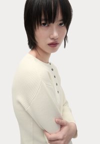 Maglione di maglia a coste color crema con scollo rotondo, caratterizzato da accenti di bottoni verde scuro e un design aderente sulle maniche e sul corpo.