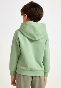 Felpa con cappuccio in cotone verde menta con cappuccio foderato, polsini e orlo a costine. Design semplice, texture morbida e vestibilità comoda, indossata sopra pantaloni grigio chiaro.