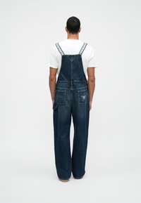 Dunkelblaue Jeanslatzhosen mit weit geschnittenen Beinen. Verfügt über verstellbare Träger und zwei Gesäßtaschen, wobei eine mit einem Sternstickereidetail versehen ist.
