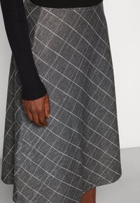 Šedá károvaná sukně s rozšířeným střihem, zdobená bílým mřížkovým vzorem. Látka působí texturovaně. Částečně viditelný černý žebrovaný dlouhý rukáv.