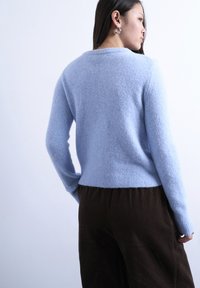 Γυναίκα με πλάτη, φορούν ανοιχτό μπλε χνουδωτό πουλόβερ και σκούρο καφέ παντελόνι, μπροστά σε απλό φωτεινό φόντο.