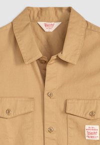 Beige overhemd met knoopsluiting en een kraag, twee borstzakken en een label. Bevat beige stof en bruine knopen, genaaid met een zachte textuur.