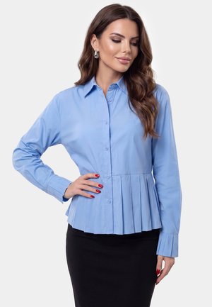 Vrouw met lang bruin haar draagt een lichtblauwe gebuttonde blouse met geplooide zoom en een zwarte rok, poseert met hand op haar heup tegen een effen achtergrond.
