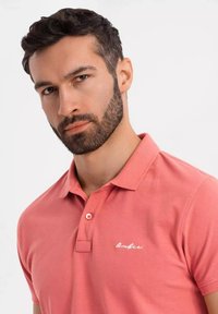 Korallinvärinen polo-paita, joka on valmistettu puuvillasta ja jossa on klassinen kaulus, kaksipainonappinen etuhalkio sekä brodeeraus vasemmassa rinnassa.