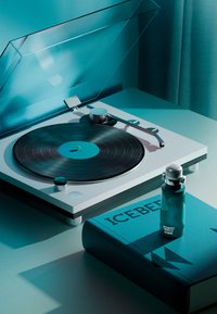 En skivspelare med en svart vinylskiva, vit bas och silverfärgad hårdvara. Bredvid den står en cyan flaska och en bok med titeln "ICEBERG." Texturerad yta.