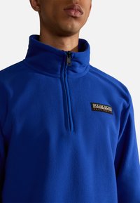 Blauwe fleecetopper met een hoge kraag, half-rits aan de voorkant en een gestructureerde stof. Kenmerkt zich door een zwart merkembleem op de borst.