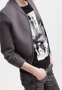 Chaqueta bomber en gris, con cremallera frontal, puños acanalados y una camiseta gráfica negra debajo con diseños fotográficos.