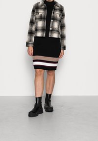 Veste courte à carreaux noir et blanc portée sur une robe noire côtelée avec des accents à rayures horizontales en beige, blanc et bordeaux. Bottines noires.