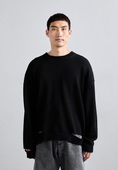 Un pull en maille noire avec un col rond, une coupe oversize et des détails de bords effilochés, associé à un jean en denim gris.