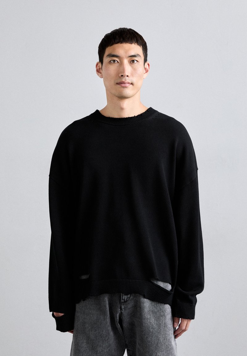 Un pull en maille noire avec un col rond, une coupe oversize et des détails de bords effilochés, associé à un jean en denim gris.