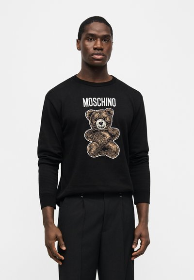 Νεαρός άνδρας που φοράει μαύρο πουλόβερ Moschino με σχέδιο αρκουδάκι, συνδυασμένο με μαύρο παντελόνι, στέκεται μπροστά σε απλό λευκό φόντο.