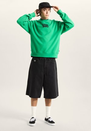Green sweatshirt with black Vans logo, black denim shorts, black cap, and black sneakers with white soles. White crew socks visible.